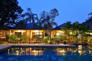 A hotel with a swimming pool, palm trees, and a well-lit exterior at dusk. - Happy Cottages Phuket (Chalong)