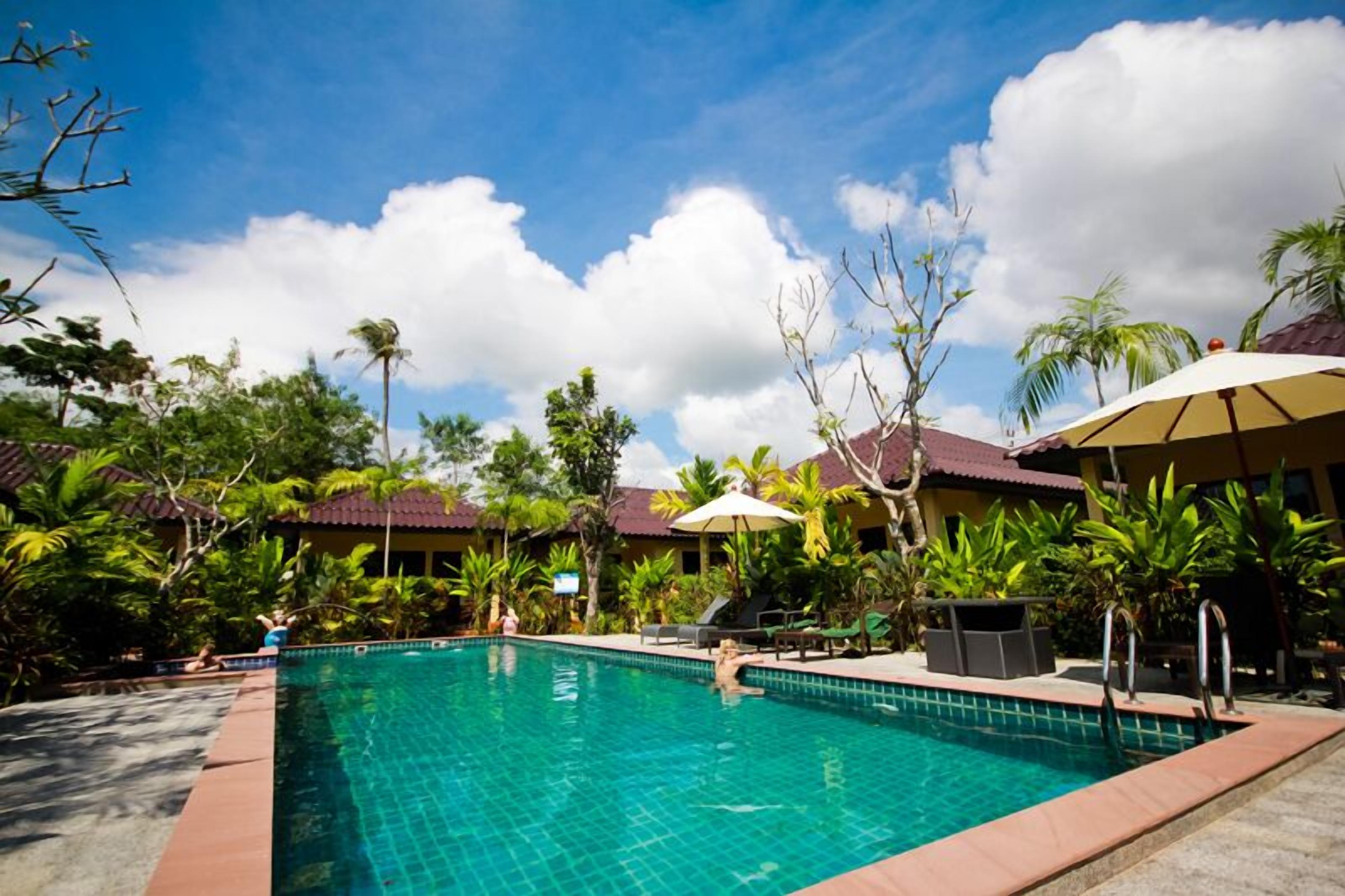 A clear swimming pool surrounded by palm trees and lounge chairs, with a building in the background.