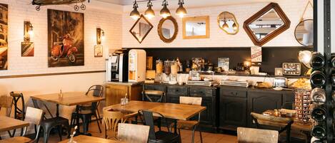 A restaurant interior with wooden tables and chairs, a kitchen area, and various wall decorations including a scooter painting and mirrors.