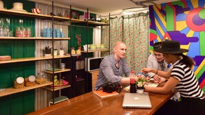 A group of people at a table with a laptop and a camera, surrounded by shelves with various kitchen items. - Vimarn Hostel (Bangkok)