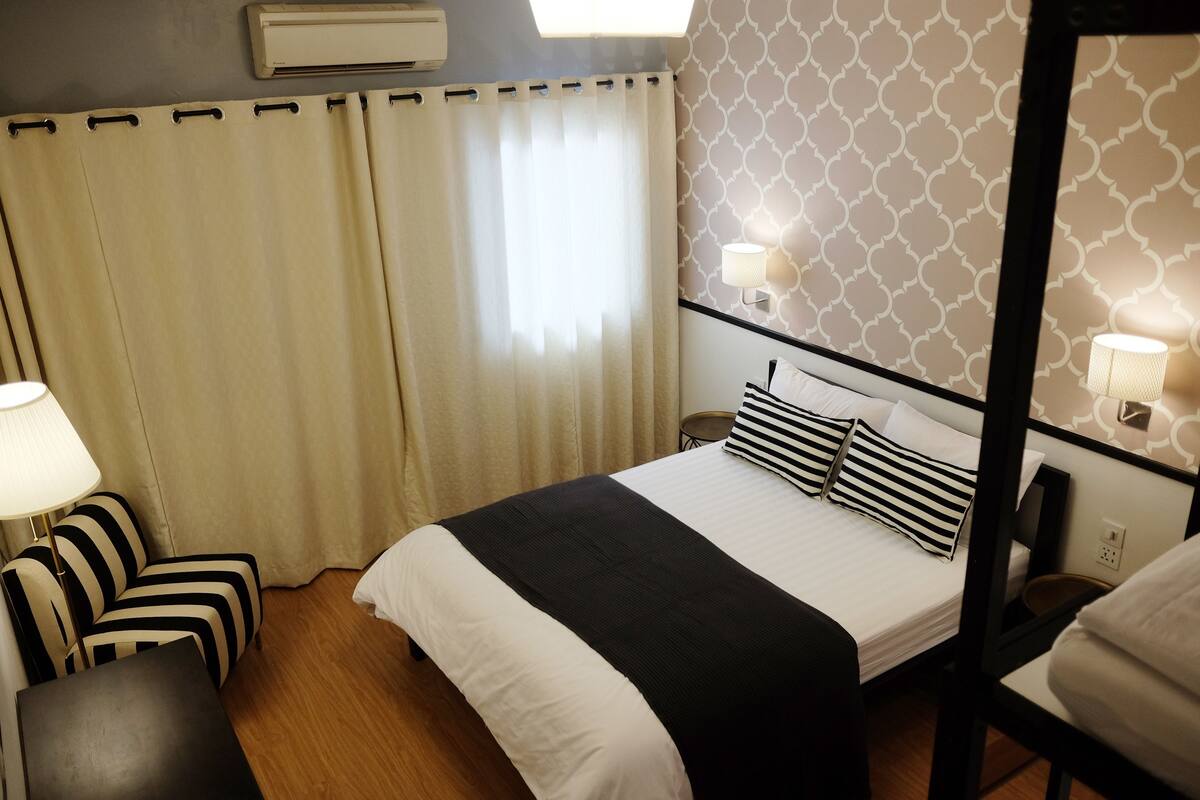 A hotel room with a bed, a bedside table, a chair, a lamp, and a window with curtains.