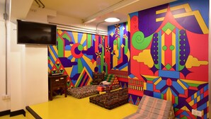 A vibrant room with colorful geometric wall art, a flat-screen TV mounted on the wall, and a small wooden side table with a remote control. - Vimarn Hostel (Bangkok)