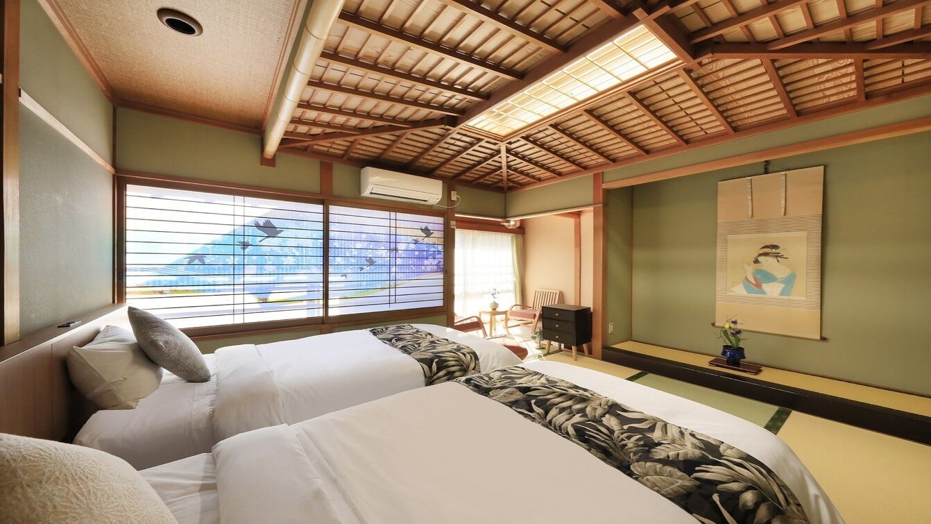 A traditional Japanese-style room with tatami flooring, a low ceiling with wooden beams, a bed with white bedding and a floral-patterned blanket, a desk with a chair, and a large window with a view of a mountain.