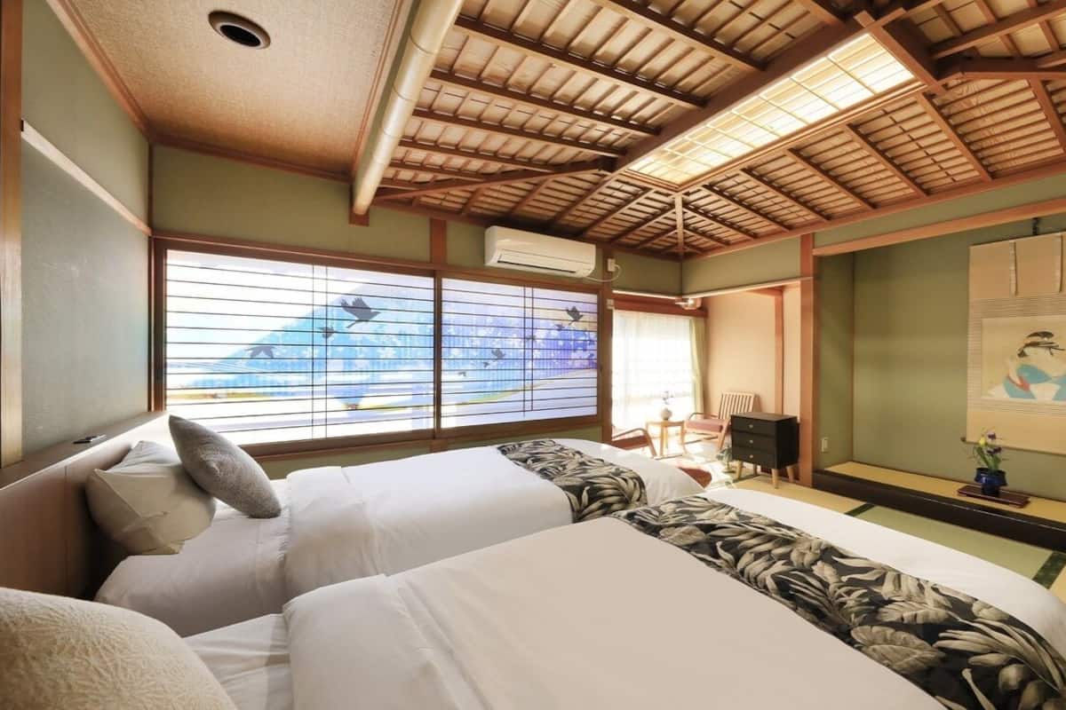 A traditional Japanese-style room with tatami flooring, a low ceiling with wooden beams, a bed with white bedding and a floral-patterned blanket, a desk with a chair, and a large window with a view of a mountain.