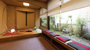 Ett traditionellt japanskt rum med tatamimattor på golvet, en låg träbänk med kuddar, en tatamimatta i mitten och utsikt över en trädgård med en stenskulptur och bambu.