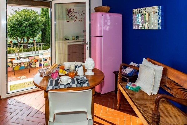 A cozy living space with a wooden table set for breakfast, a pink refrigerator, a wicker sofa, and a view of a garden through a glass door. - Il Mirto Bed and Breakfast (Imperia)