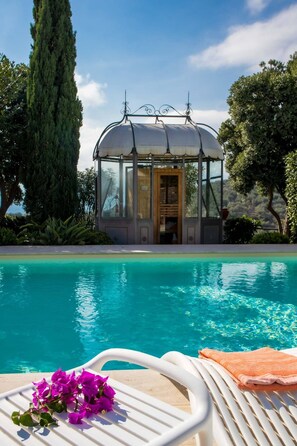 A glass-enclosed structure with a door, a pool with clear blue water, and a white lounge chair with a towel. - Il Mirto Bed and Breakfast (Imperia)