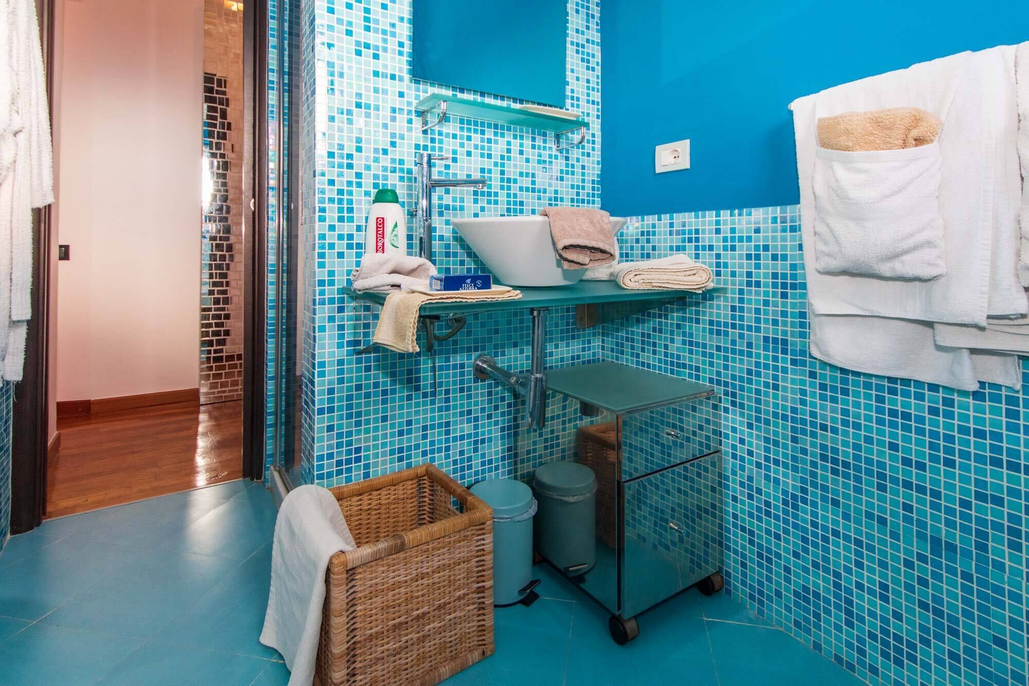 Un baño con mosaicos azules, un perchero blanco, una cesta de mimbre y un carrito pequeño.