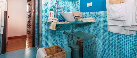 A bathroom with blue mosaic tiles, a white towel rack, a wicker basket, and a small cart.