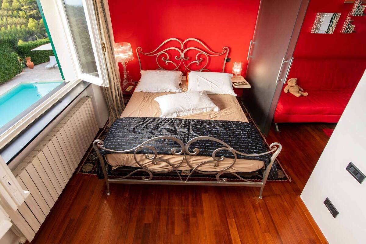 A bedroom with a bed, a red wall, a window, and a red sofa.