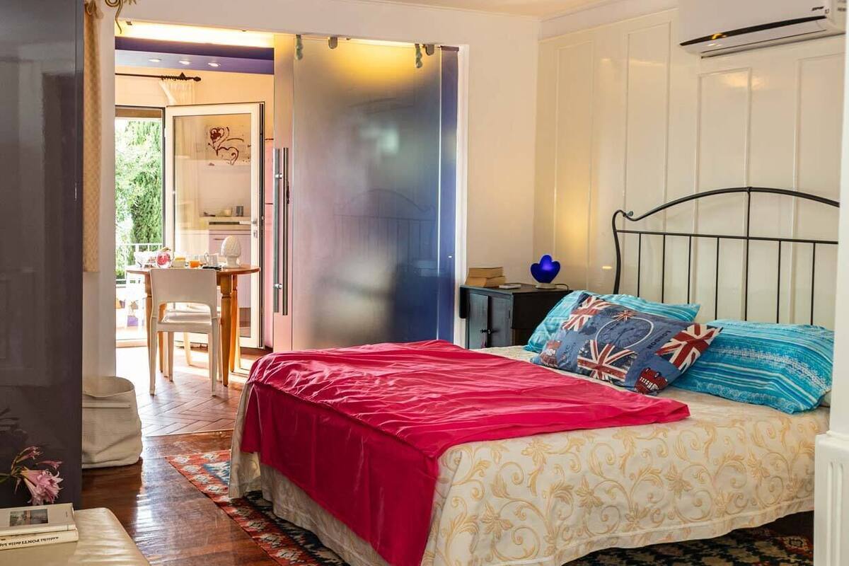A bedroom with a bed, a chair, a small table, and a door leading to another room.