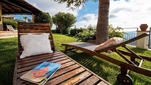 Una terraza de madera con una silla de descanso y una revista, rodeada de follaje y un cielo despejado.