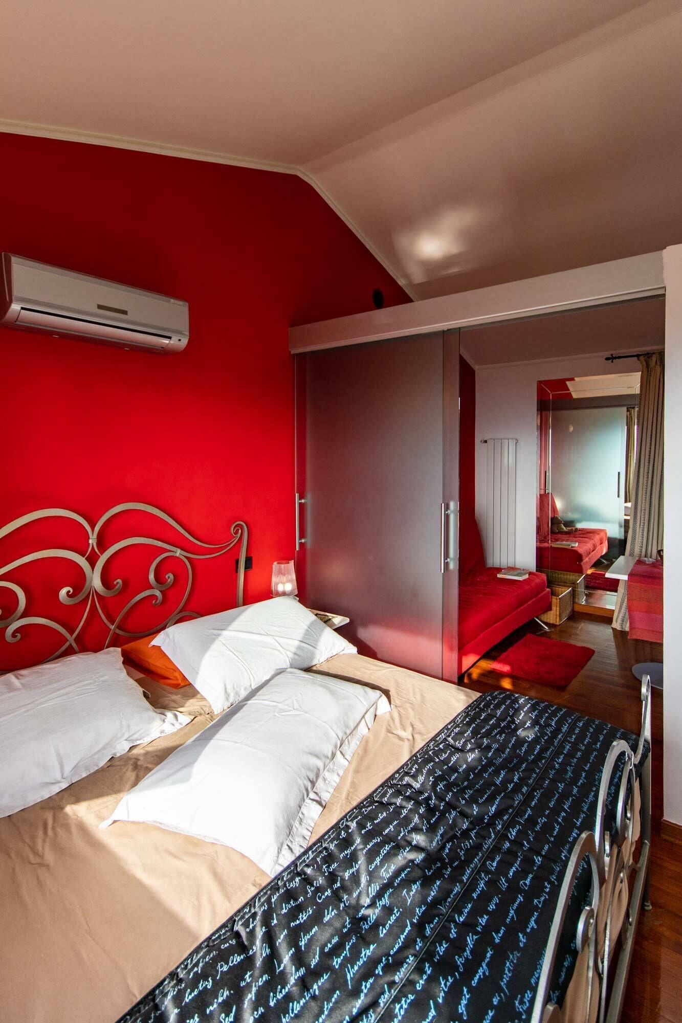 Camera da letto con una parete rossa, un letto con biancheria da letto bianco e arancione, una coperta a righe nere e un armadio rosso per abiti.