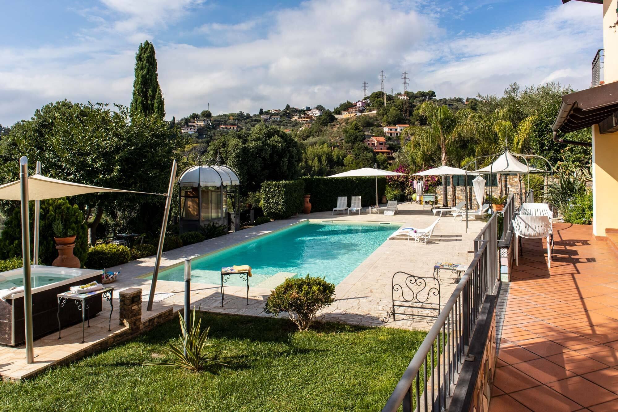 A swimming pool with lounge chairs and umbrellas, surrounded by greenery and a view of a hillside with houses.