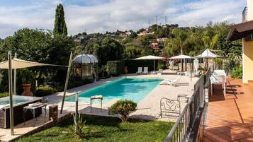 A swimming pool with lounge chairs and umbrellas, surrounded by greenery and a view of a hillside with houses.