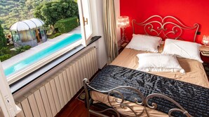 Un dormitorio con cama, ventana con vista a una piscina y follaje, pared roja y lámpara.