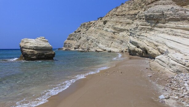 Una playa costera con aguas azules cristalinas, orilla de arena y formaciones rocosas en capas.