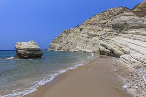 A coastal beach with clear blue water, sandy shore, and layered rock formations. - Fourtounis Hotel (Kos)