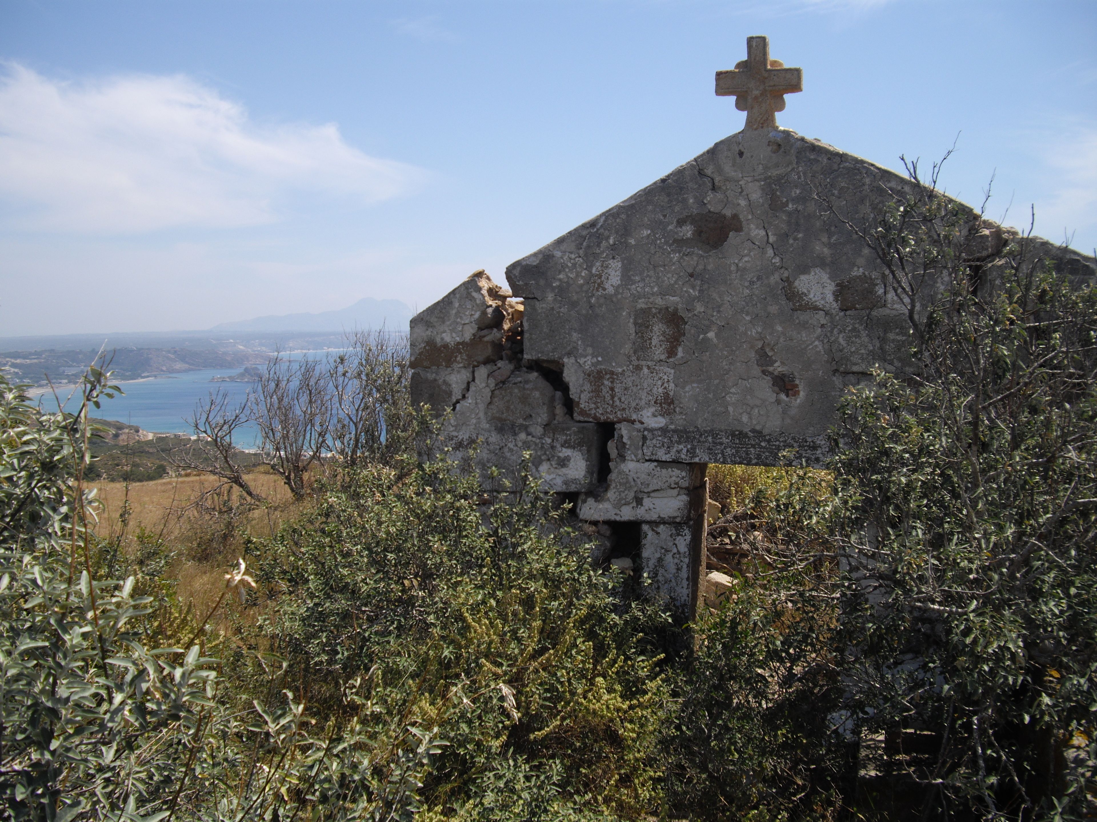 Una cruz de piedra erosionada en la cima de una estructura en ruinas, rodeada de vegetación densa.