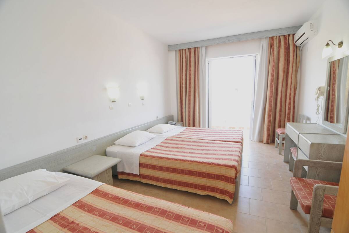 A hotel room with two beds, a bench, a desk, a mirror, and a window with curtains.