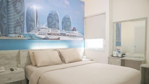 Quarto de hotel com uma cama grande, mesa de cabeceira com telefone e controle remoto, banheiro com espelho e um mural na parede retratando um navio de cruzeiro e edifícios.