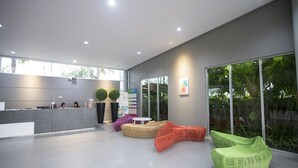 Uma área de recepção moderna com balcão, espaço de estar com cadeiras coloridas e grandes janelas com vista para o verde.