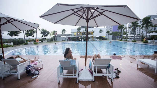 Uma área à beira da piscina com espreguiçadeiras e guarda-sóis, uma piscina e palmeiras ao fundo.