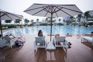 A poolside area with lounge chairs and umbrellas, a swimming pool, and palm trees in the background.