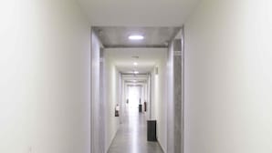 Um corredor comprido e estreito, paredes brancas e chão cinzento, que leva a uma porta no final.
