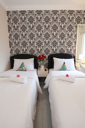 A hotel room with two beds, each with a pillow and a towel. There is a nightstand with a phone and a vase with flowers. The walls have a patterned wallpaper.