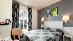 A hotel room with a bed, two bedside lamps, a window with curtains, a chair, a small table with a red book, and a wall art piece. - Hôtel Miramar (Paris)