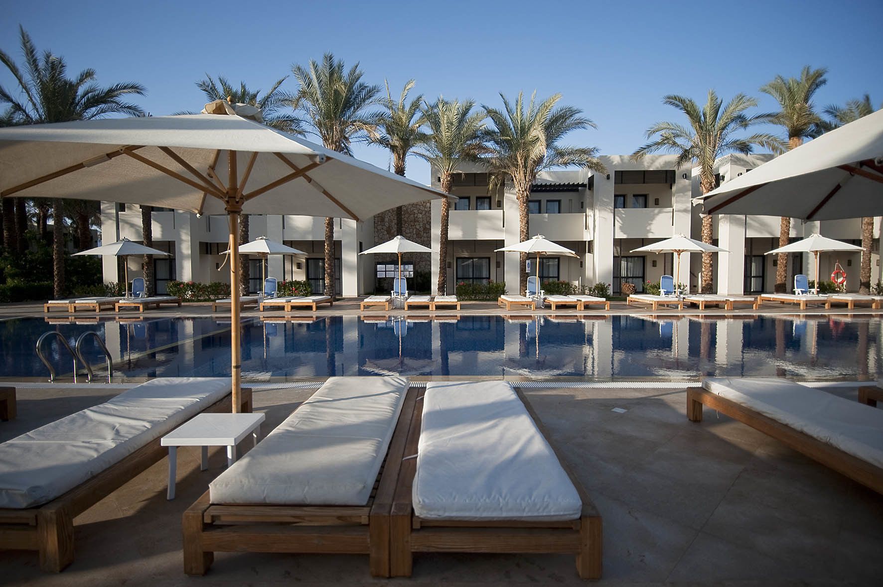 A pool area with lounge chairs and umbrellas, surrounded by palm trees and a modern building.