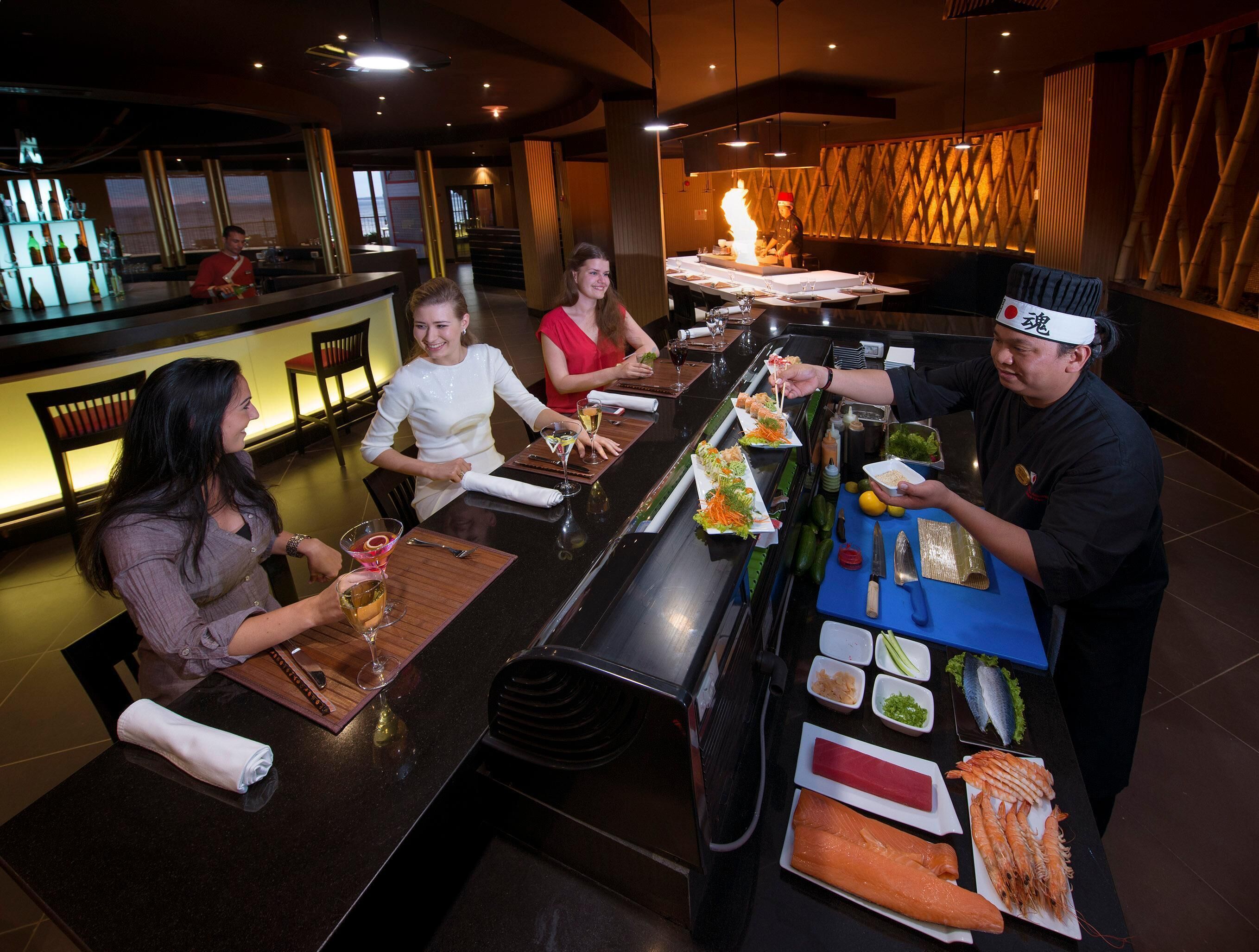 A sushi bar with a chef preparing dishes, a bar area with bottles, and a seating area with tables and chairs.