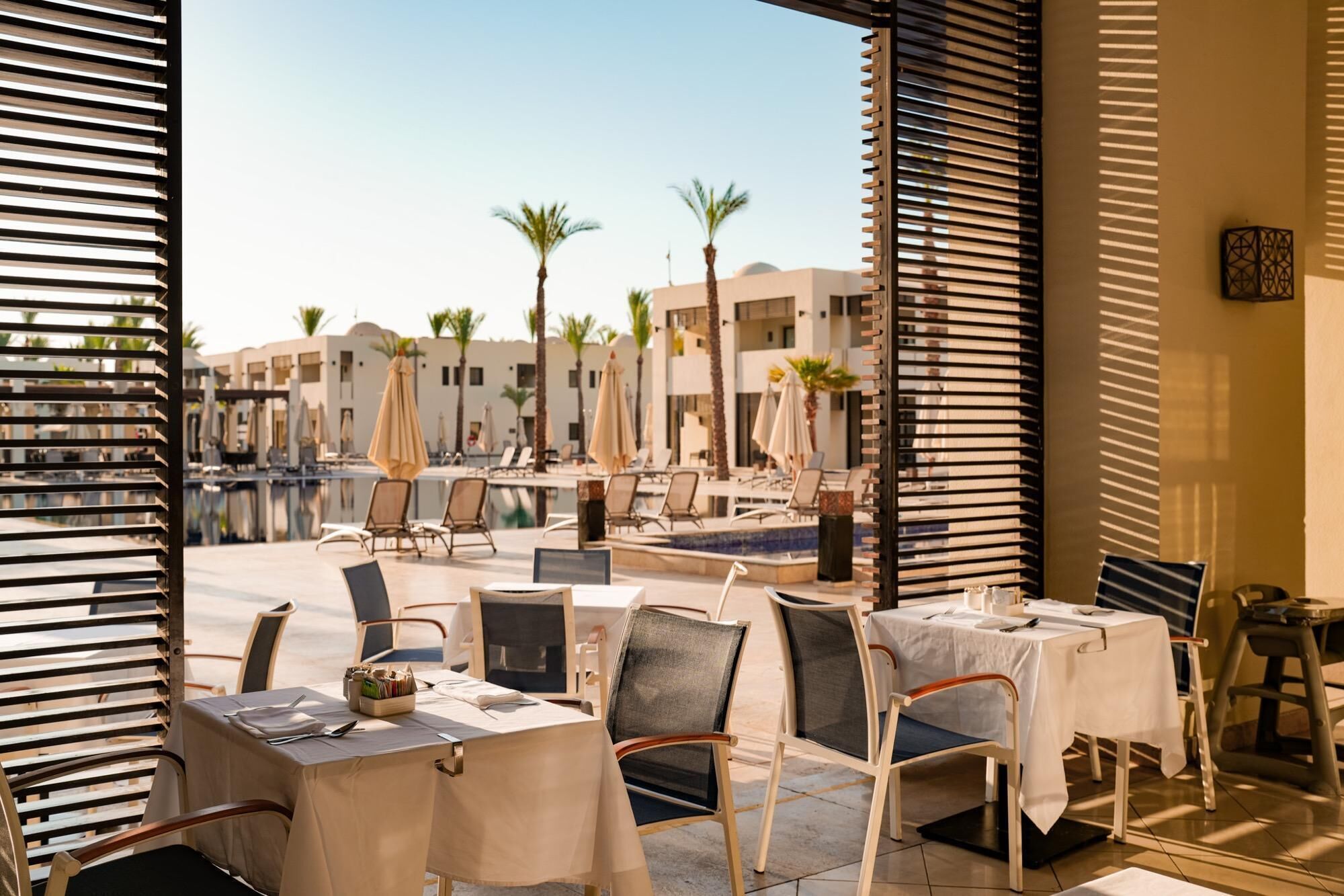 An outdoor dining area with tables and chairs, a pool, and palm trees in the background.