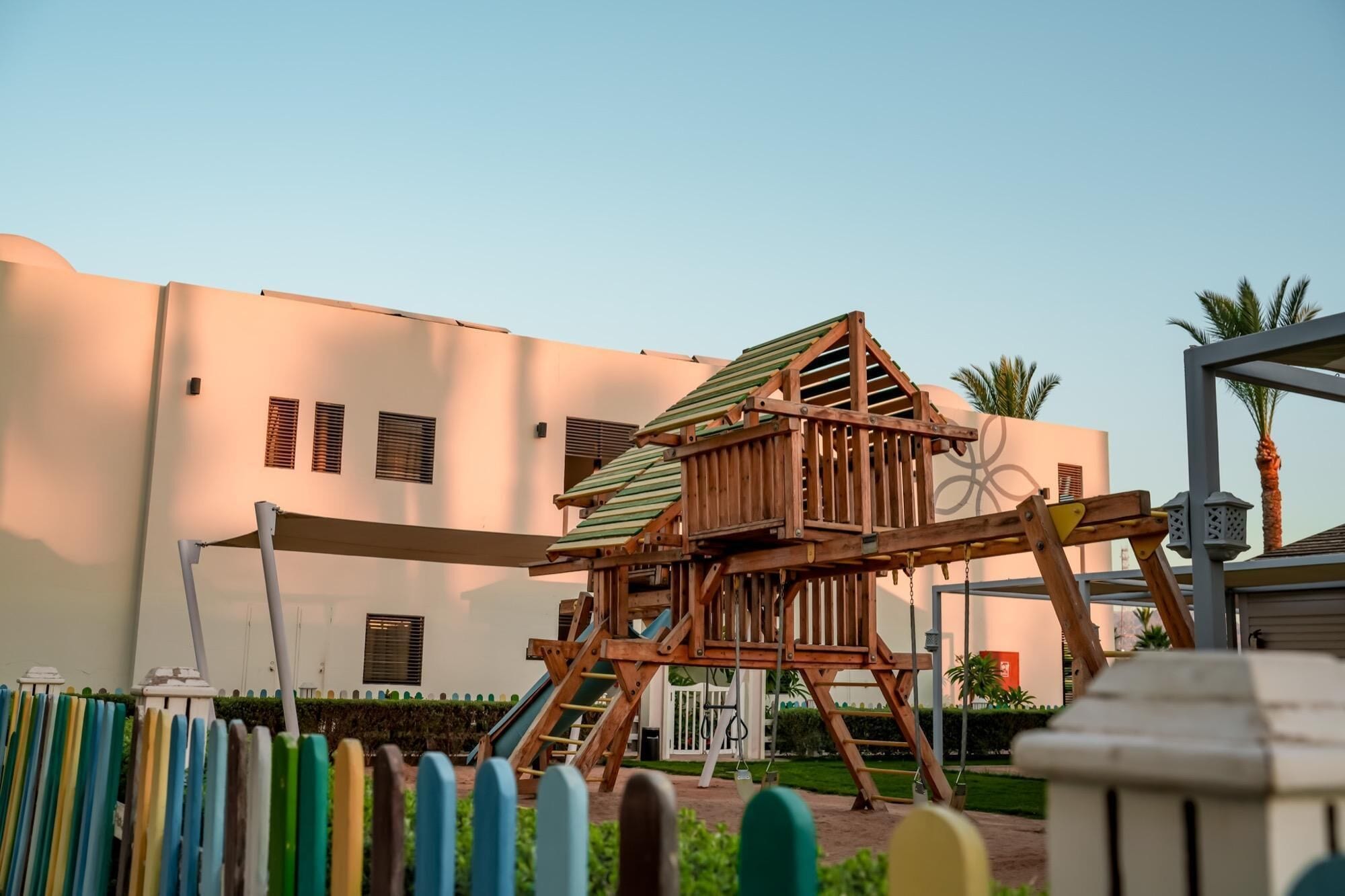 A playground with a wooden swing set and a slide, set against a building with multiple windows and a palm tree in the background.