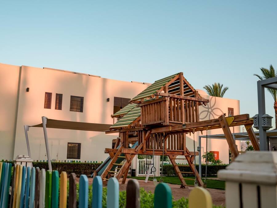 A playground with a wooden swing set and a slide, set against a building with multiple windows and a palm tree in the background.