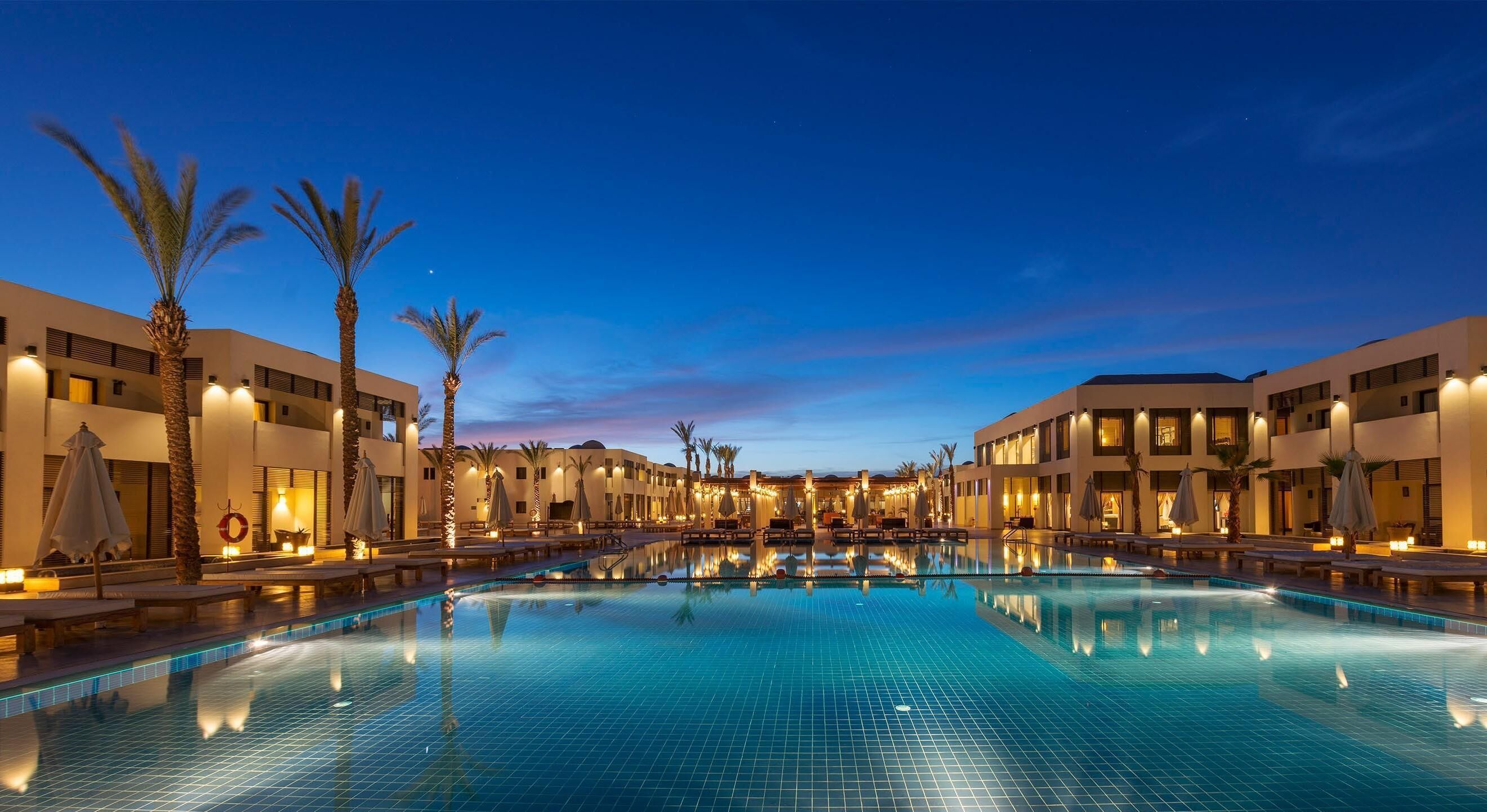 A hotel pool area with multiple lounge chairs and umbrellas, surrounded by palm trees and modern buildings.