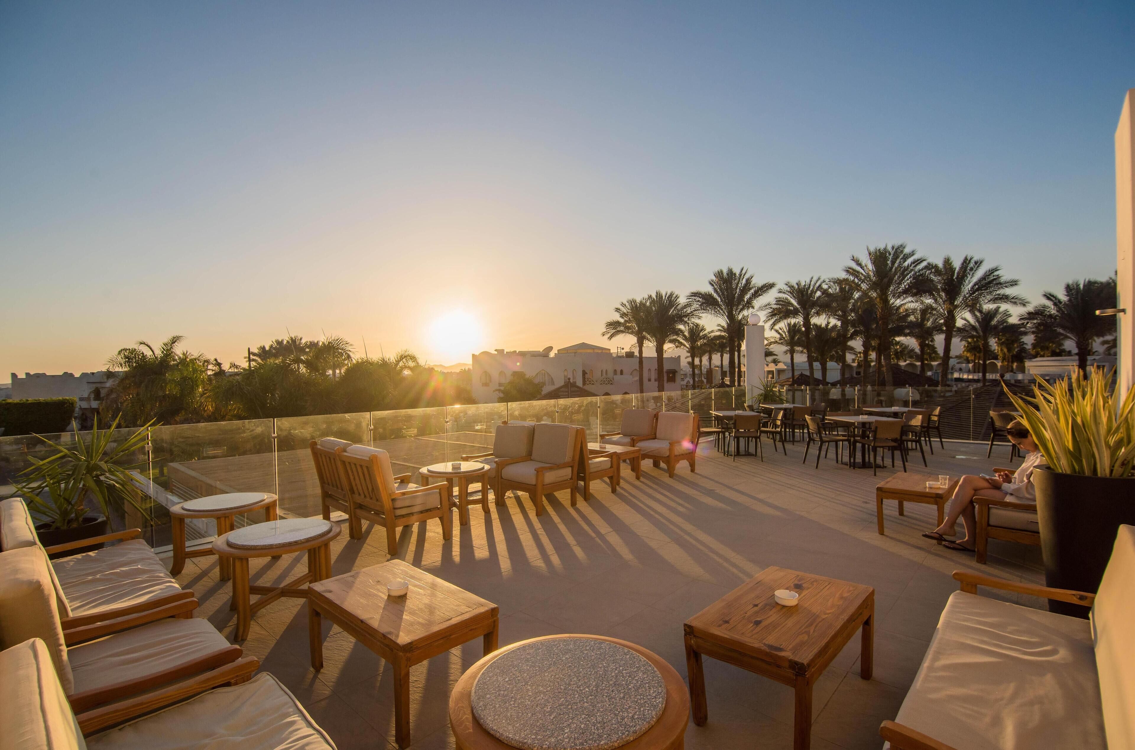A rooftop terrace with wooden furniture, palm trees, and a sunset view.