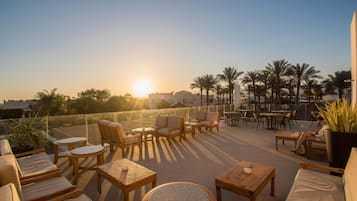 A rooftop terrace with wooden furniture, palm trees, and a sunset view.