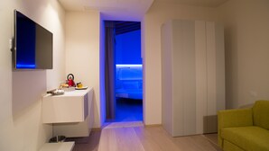 Una habitación de hotel moderna con televisor de pantalla plana, una kitchenette pequeña, una cama con dosel iluminado en azul y un sofá verde.