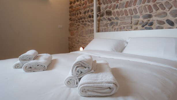 Una cama bien hecha con toallas blancas enrolladas y una pared de piedra al fondo.