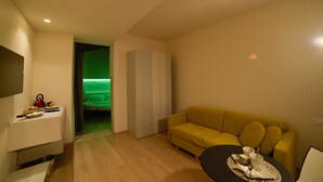 Una sala moderna con un sofá amarillo, una mesita con una bandeja, un armario blanco y una habitación iluminada en verde al fondo.