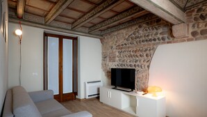 Una sala de estar con una pared de piedra, un televisor de pantalla plana sobre un mueble blanco, un sofá gris y una lámpara en una mesita.