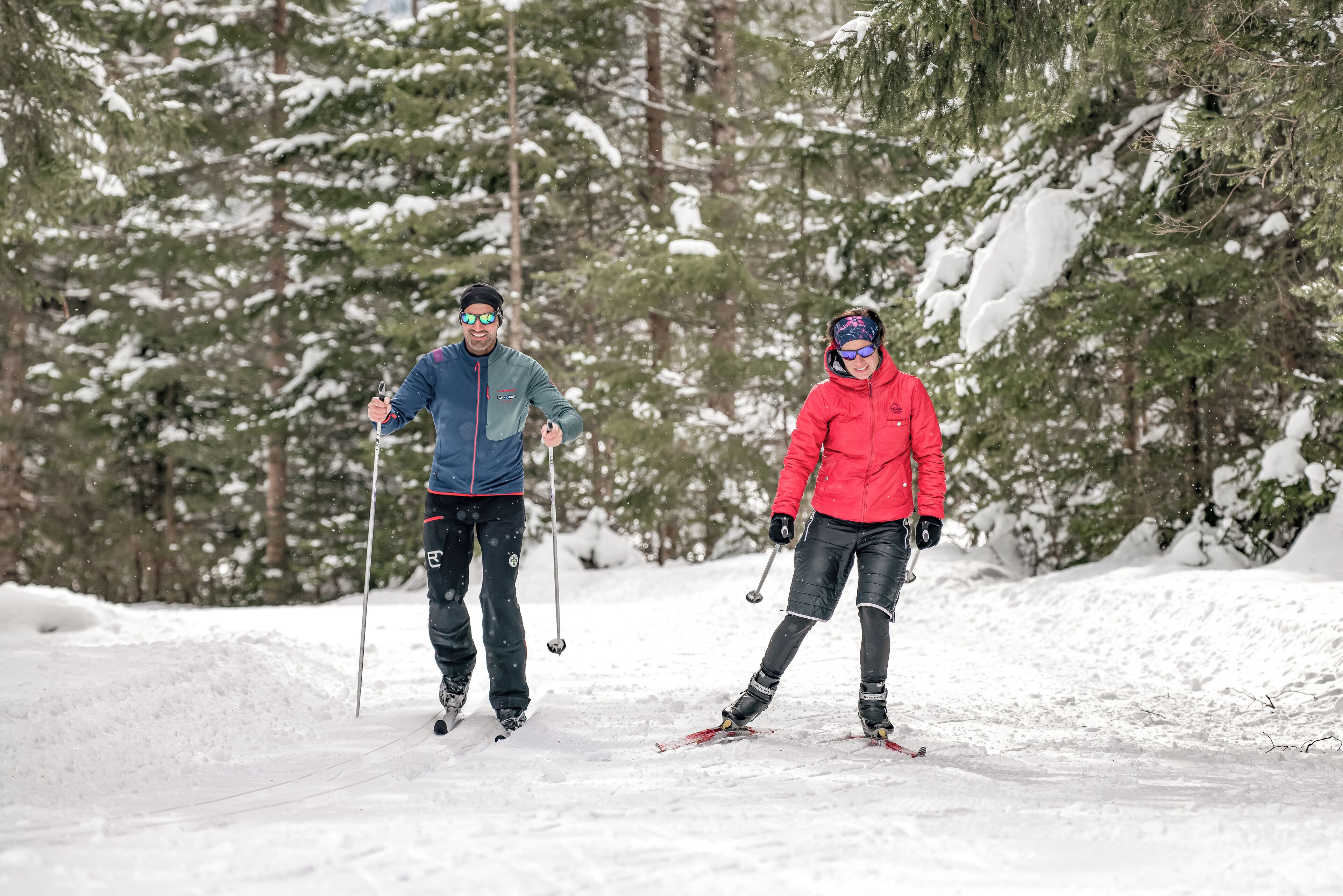 Two people cross-country skiing in a snowy forest.