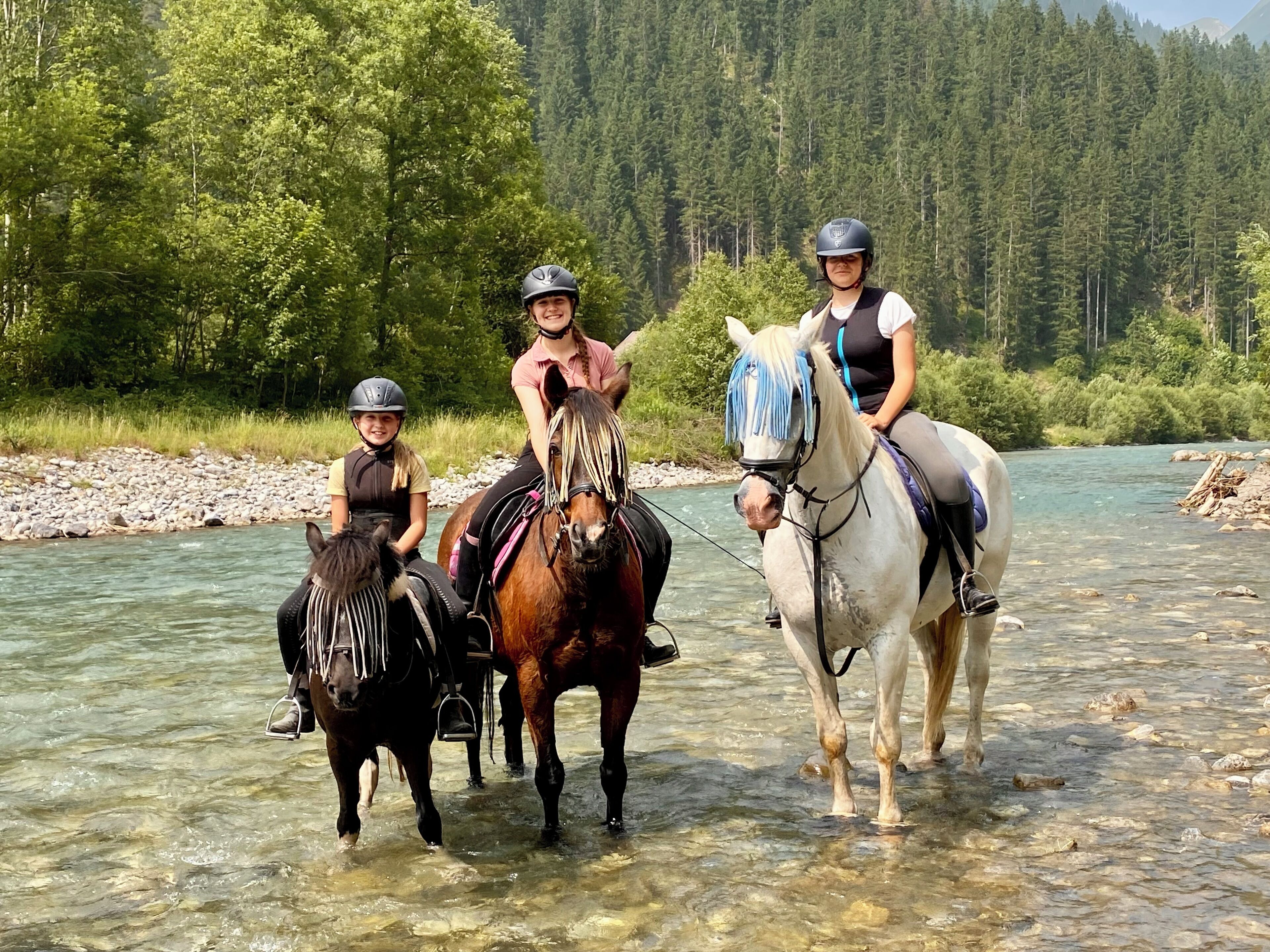 Tres personas montando a caballo por un río poco profundo con un bosque al fondo.