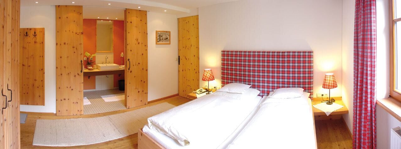 A bedroom with a large bed, wooden wardrobe, a bathroom with a sink and mirror, and two bedside lamps.