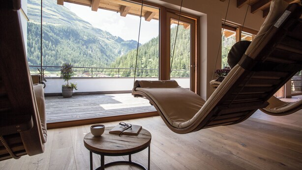 A room with a wooden floor, a hanging chair, a small table with a bowl and a book, and large windows offering a mountain view.