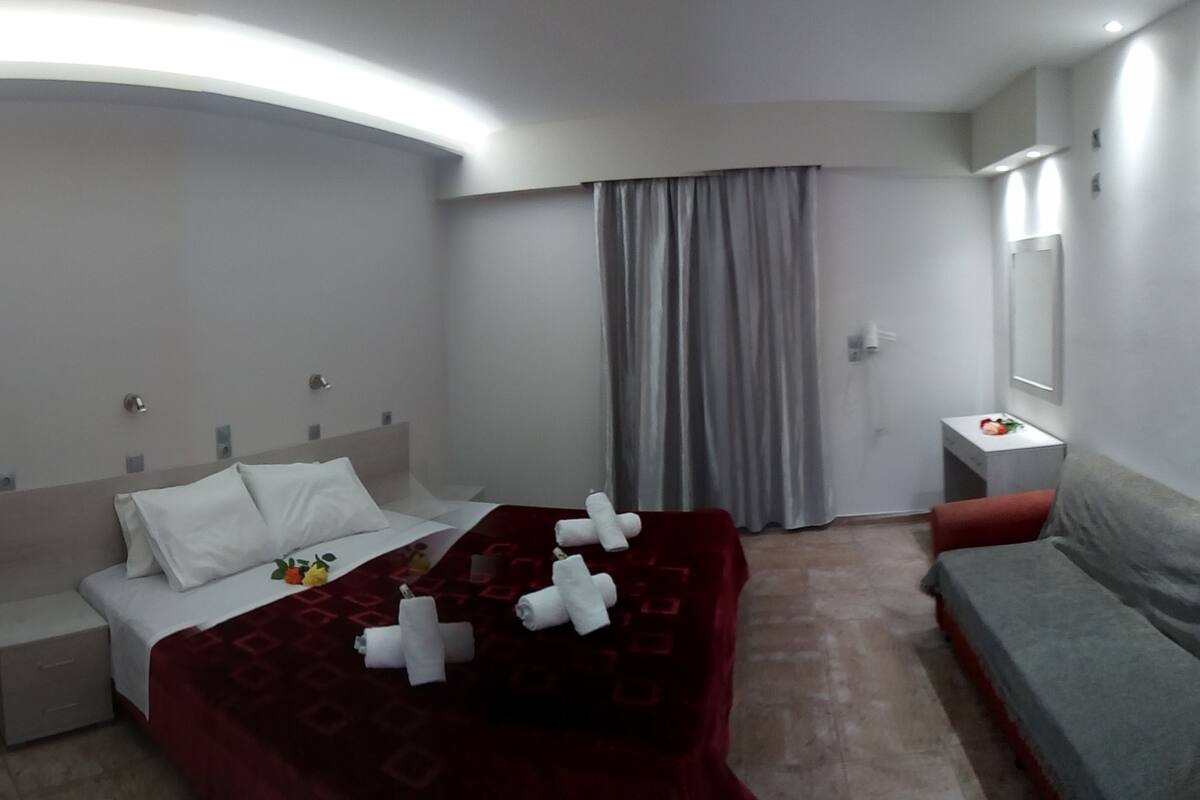 A hotel room with a bed, a bedside table, a nightstand, a sofa, a mirror, and a window with curtains.