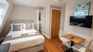 A modern hotel room with a neatly made bed, a small wooden table with a chair, a TV mounted on the wall, and a door leading to another room. - Imperial Court Studios (London)