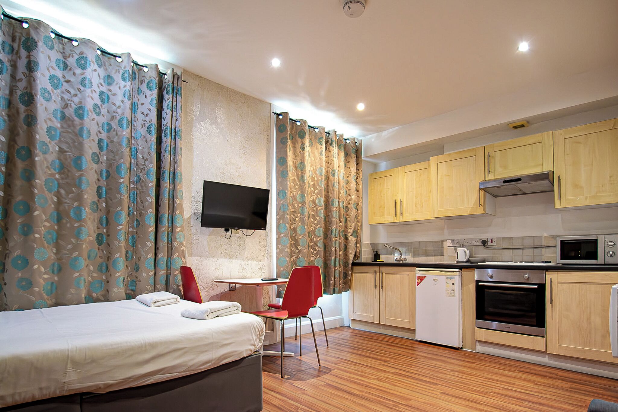 Habitación de hotel compacta con cama, un pequeño escritorio, una silla, kitchenette con microondas, horno y refrigerador, y televisor montado en la pared.
